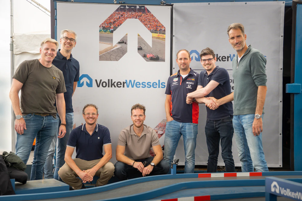 Een groep van acht mensen poseert voor een VolkerWessels-banner, met een kleine racebaan vooraan.