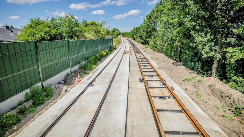 Spoorwegconstructie met twee sporen op een betonnen ondergrond, linkerzijde groene geluidswal en bomen aan de rechterkant; in de verte een oranje-witte veiligheidspaal.