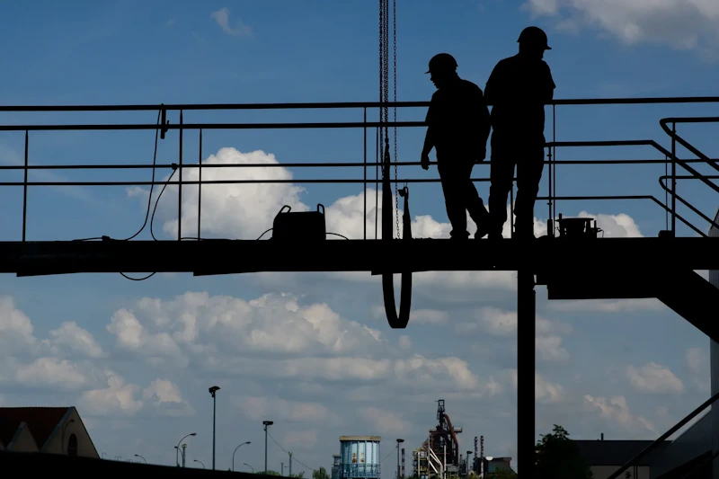 Twee silhouetten van arbeiders met helmen staan op een hangende werkbrug, tegen een blauwe hemel met wolken en een industriële installatie op de achtergrond