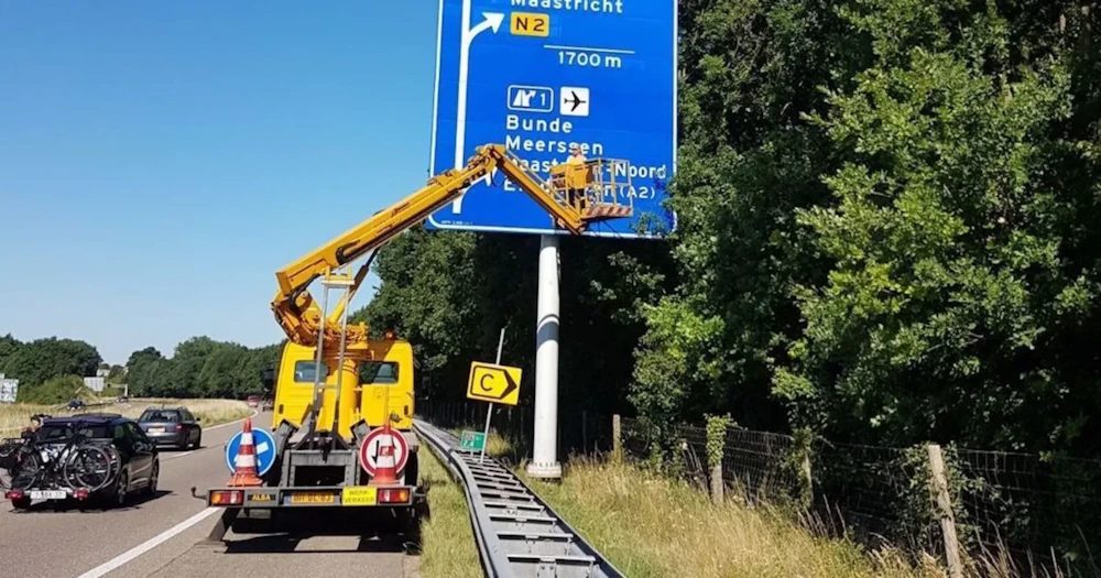 Geel hoogwerker op een vrachtwagen werkt aan een grote blauwe wegwijzer langs een landweg, met auto en bomen op de achtergrond.